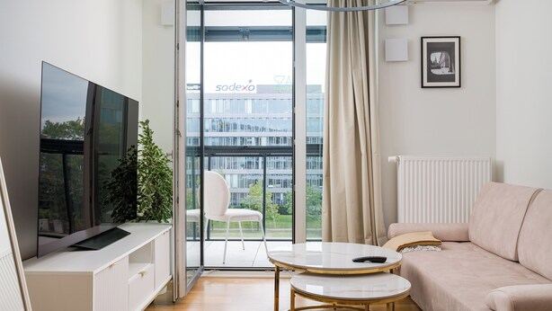 Ein modernes Wohnzimmer mit Flachbildfernseher, Sofa, Couchtisch und einem Balkon mit Blick auf ein Gebäude.