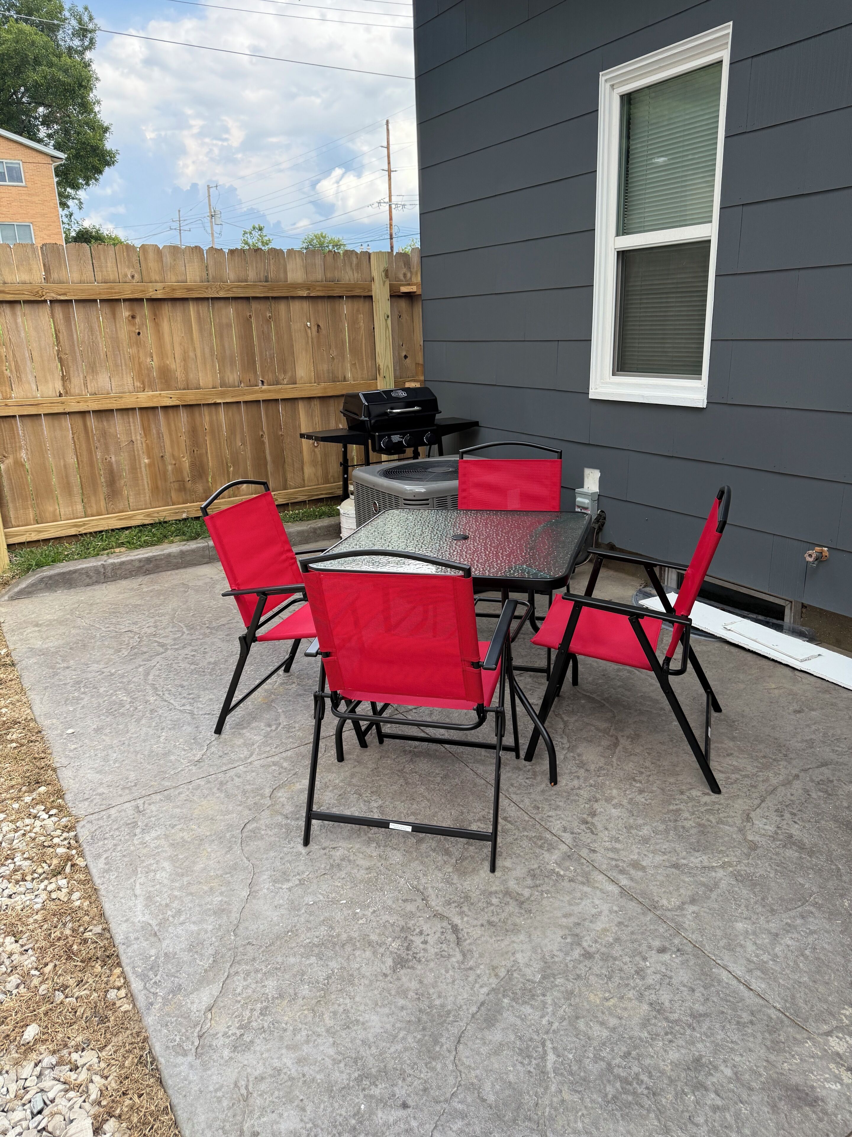 A patio with a table and chairs, a grill, and a wooden fence in the background.