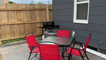 A patio with a table and chairs, a grill, and a wooden fence in the background.