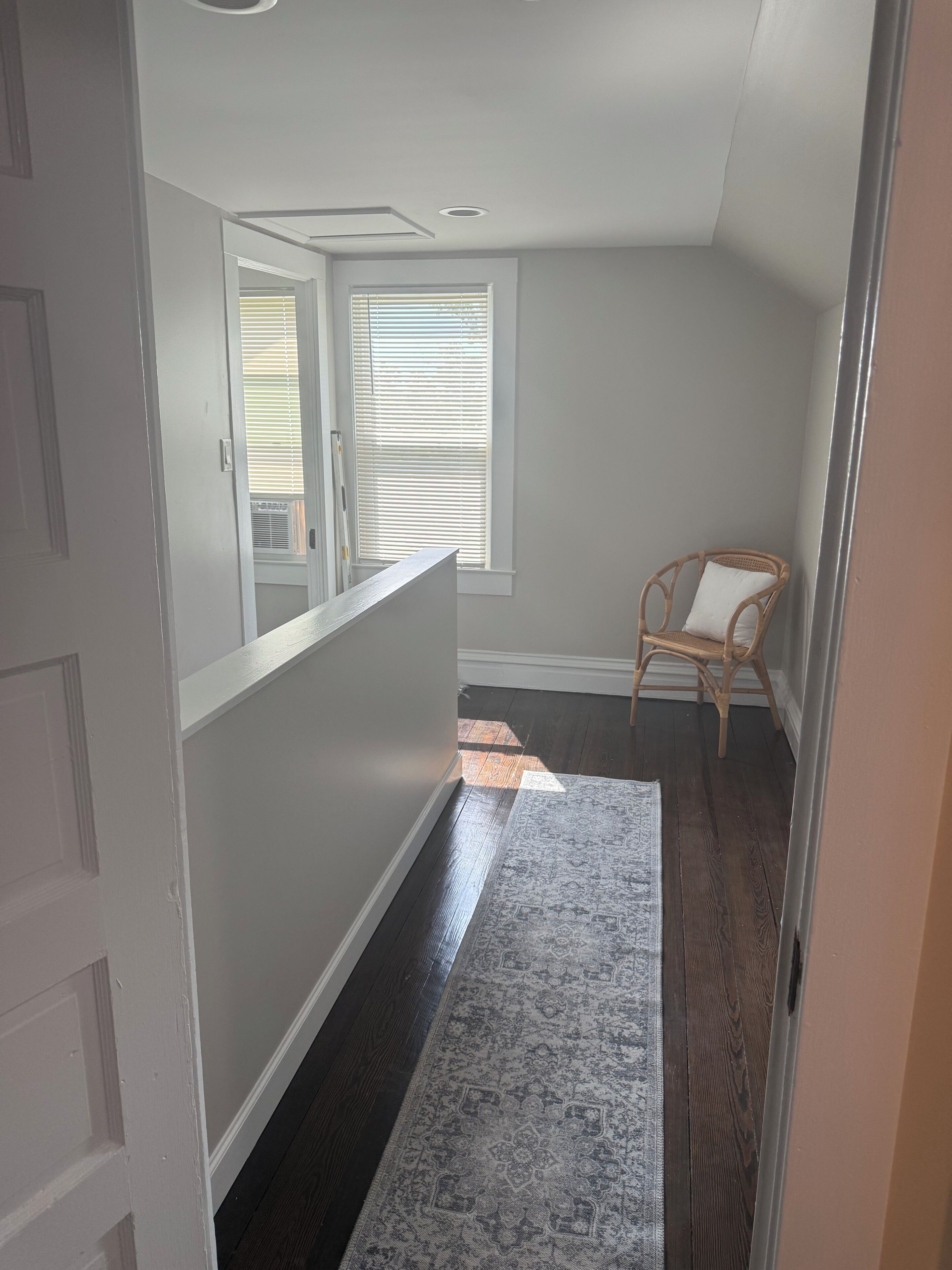 A hallway with a dark wooden floor, a patterned rug, a wicker chair, and a window with blinds.