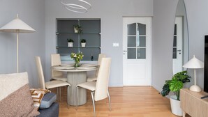 A modern living room with a round dining table, beige chairs, a sofa with patterned cushions, a floor lamp, a potted plant, and a large mirror.