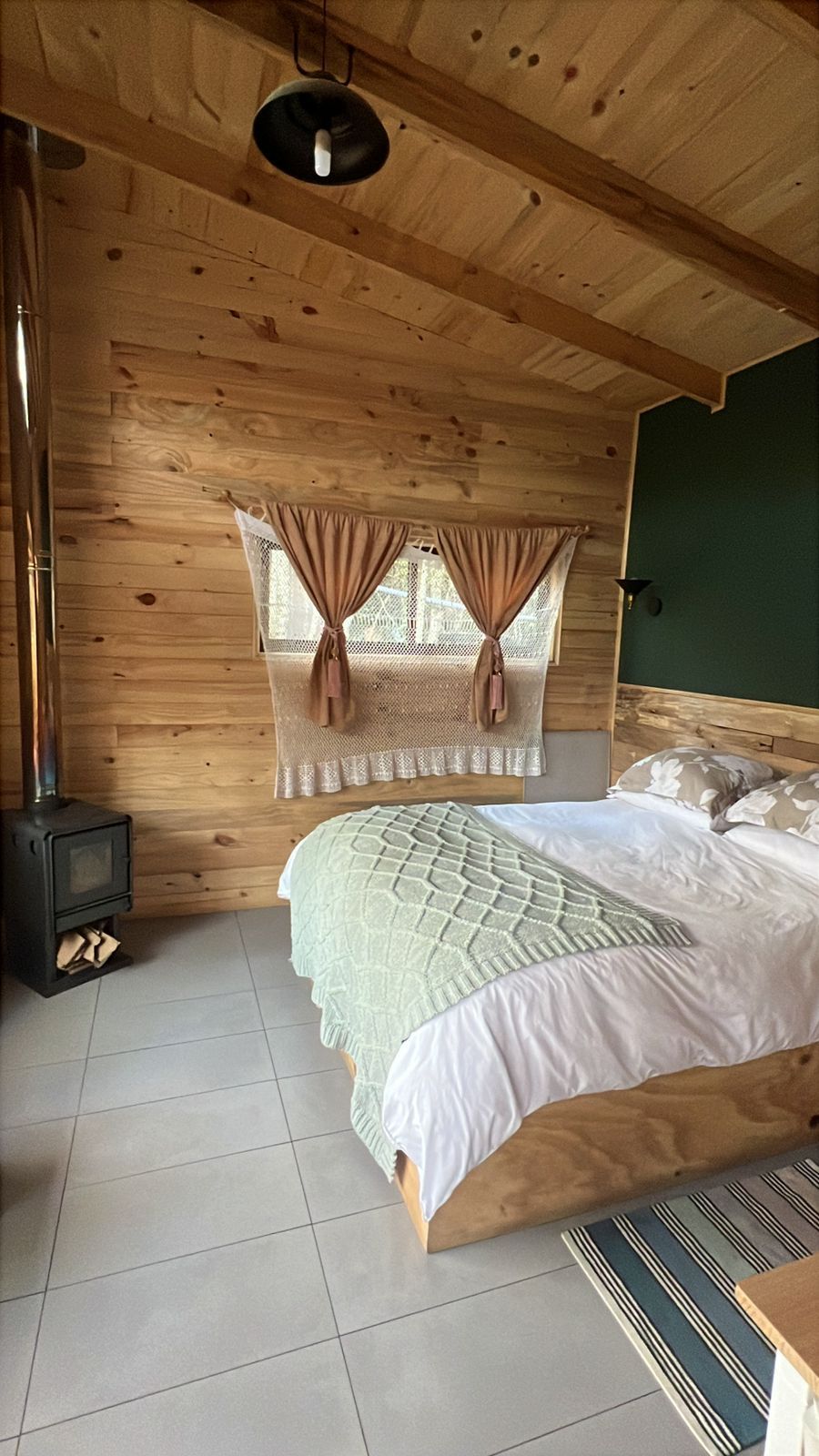 A wooden cabin bedroom with a bed, a window with curtains, a heater, and a green wall.