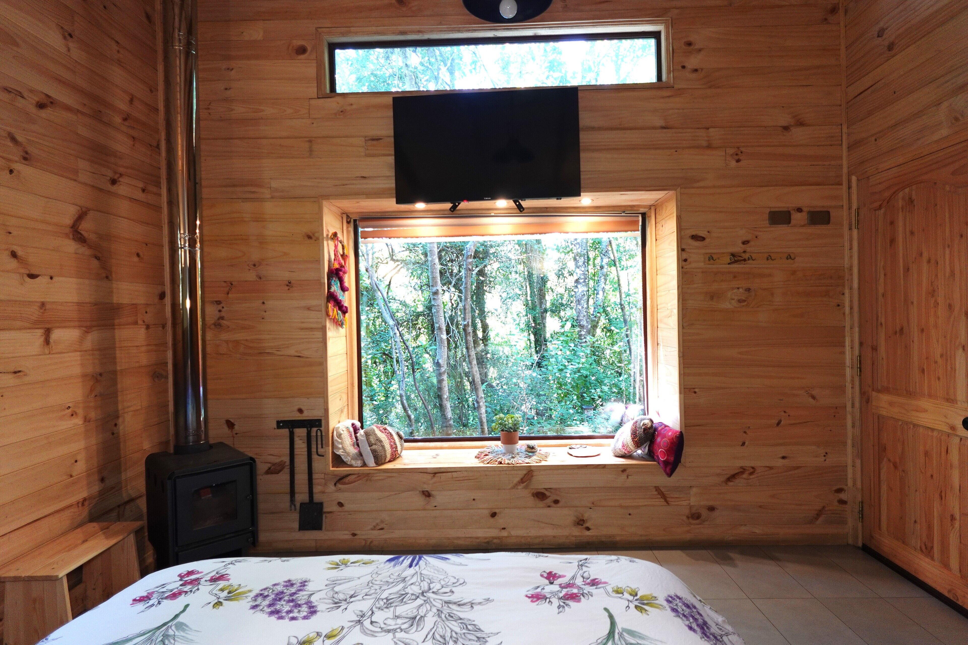A cozy cabin room with a bed, a window with a view of trees, a fireplace, and a television mounted above the window.