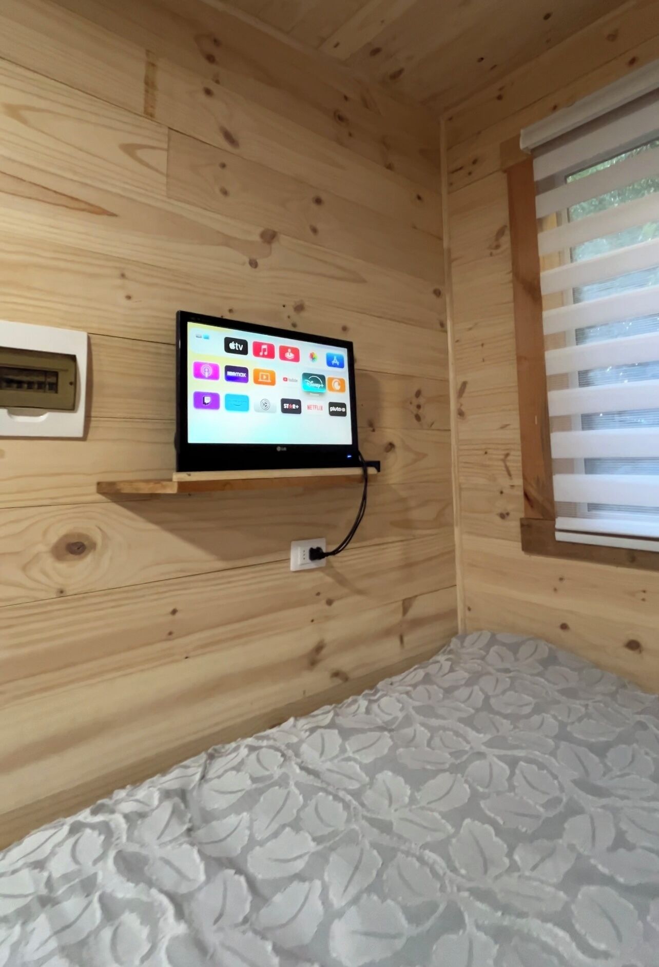A wooden cabin room with a bed, a mounted television displaying various streaming apps, and a window with blinds.