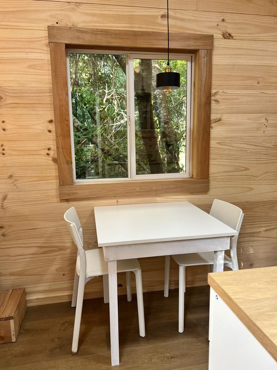 A wooden interior with a white table and chairs, a window with a view of trees, and a hanging light fixture.