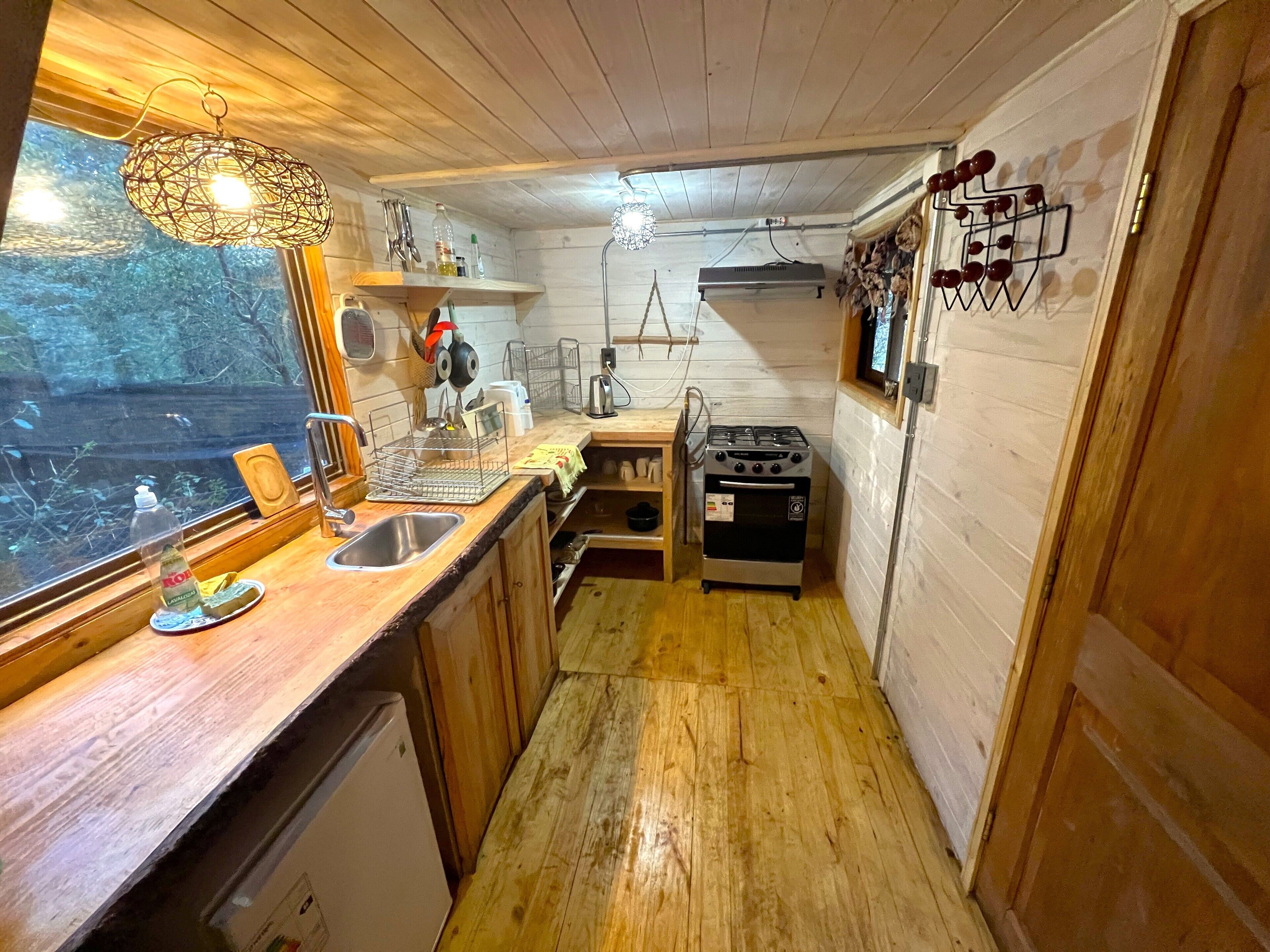 A compact kitchen with wooden cabinets, a sink, a stove, and a window overlooking a forested area.
