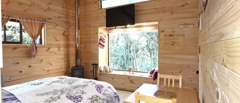 A wooden cabin room with a bed, a table, and a window overlooking a forest.