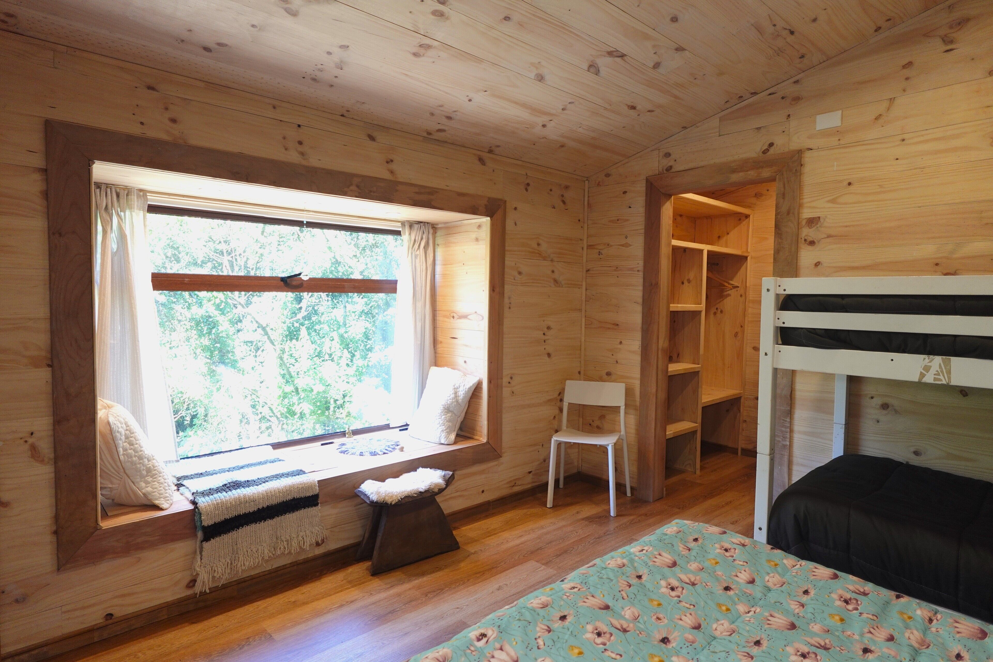 Une chambre dans un chalet en bois, avec un lit superposé, une fenêtre avec des rideaux, une chaise et une petite table recouverte d’une couverture.