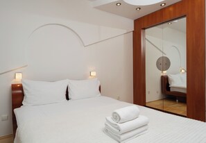 Een net opgemaakt bed met witte lakens en een stapel opgevouwen handdoeken, een houten hoofdbord en een grote spiegel aan de muur.