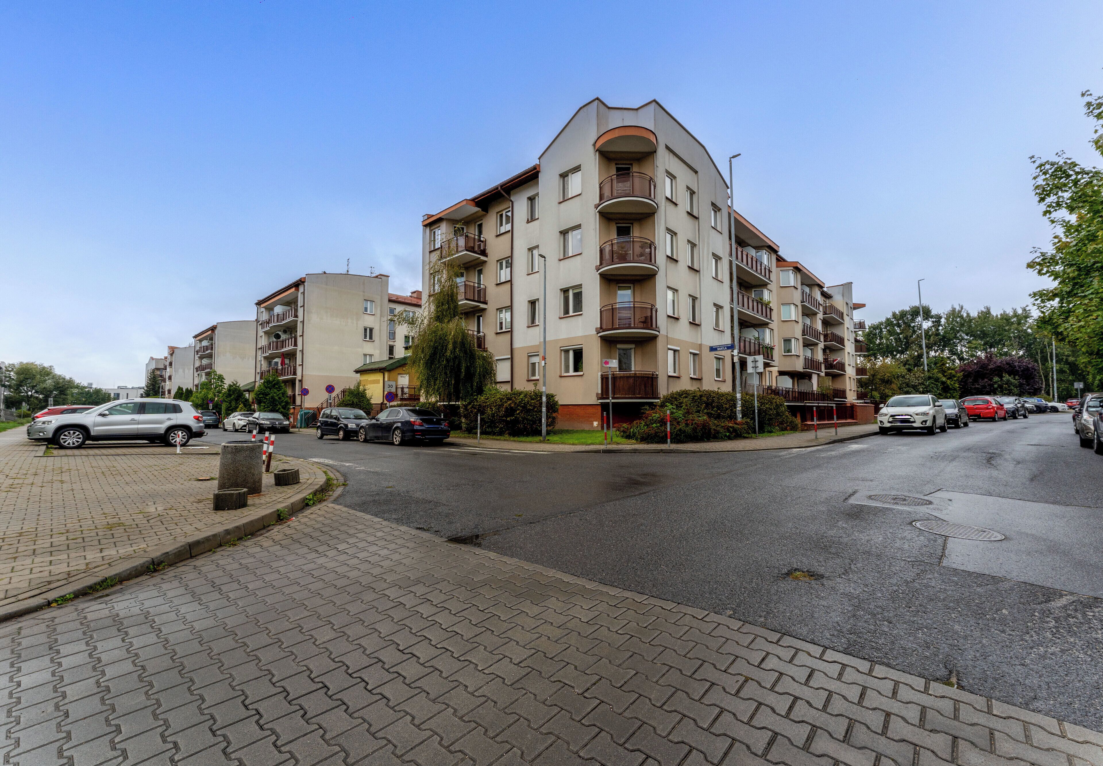 Un edificio residencial de varios pisos con balcones, una acera pavimentada y coches aparcados.