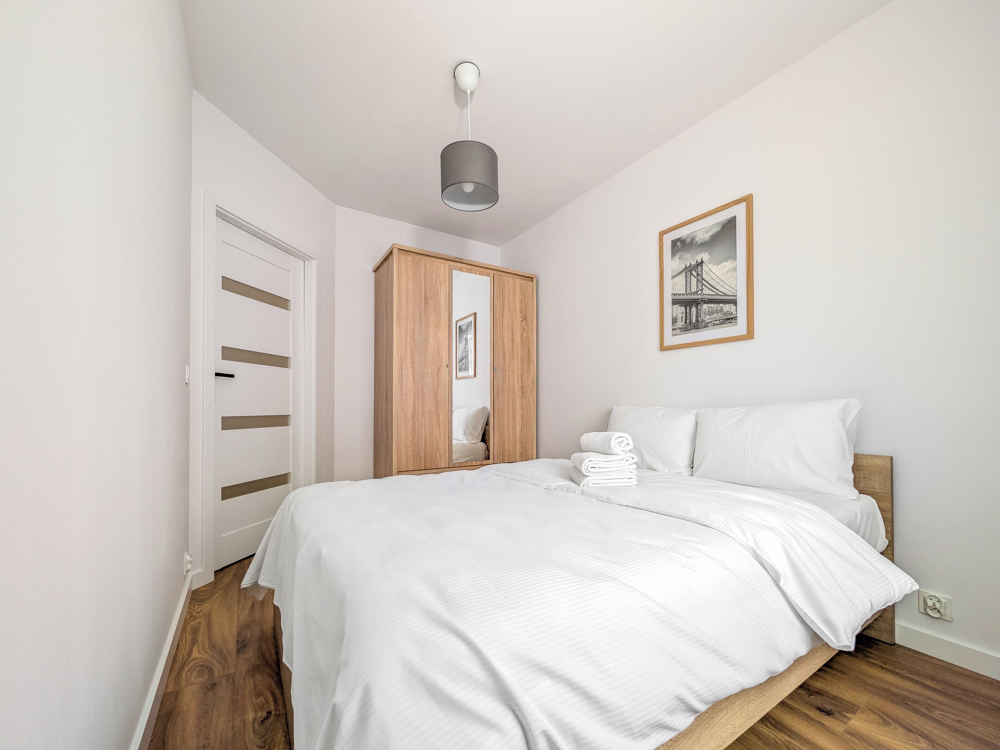 Quarto com cama, guarda-roupa de madeira, espelho, quadro emoldurado e luminária no teto.