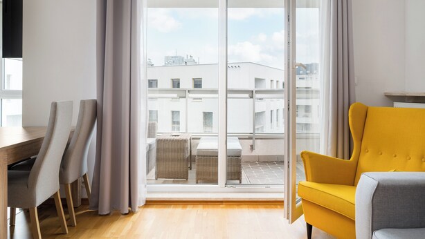 Un salon moderne avec une porte-fenêtre coulissante donnant sur un balcon, un fauteuil jaune et un coin repas avec des chaises.