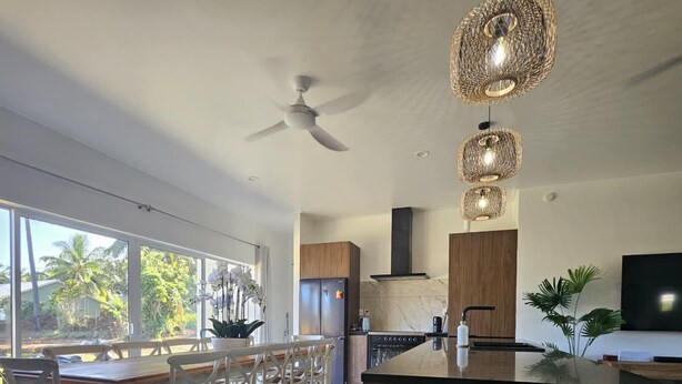 Une cuisine moderne dotée d’un grand îlot central, de meubles en bois et d’un four encastré. On y trouve également un coin repas avec des chaises blanches et une grande fenêtre donnant sur un paysage tropical.
