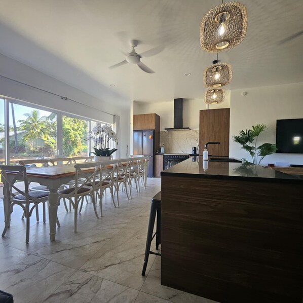 A modern kitchen with a large island, wooden cabinets, and a built-in oven. There is a dining area with white chairs and a large window overlooking a tropical landscape.