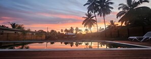 A swimming pool with a wooden deck, surrounded by palm trees and a sunset view.