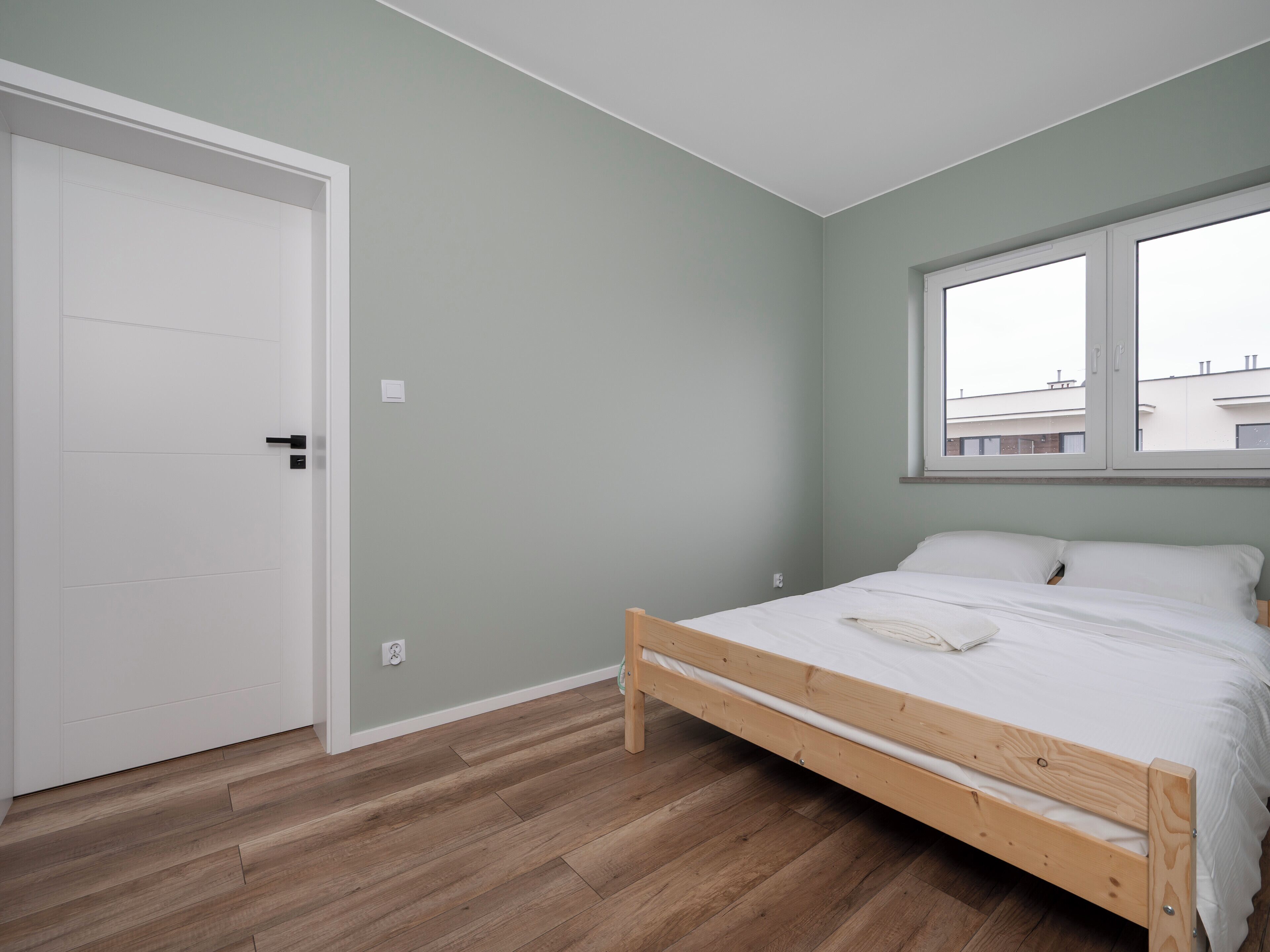 A bedroom with a wooden bed, a white door, and a window with a view of buildings.