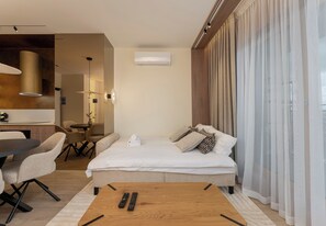 Habitación de hotel moderna con cama, una mesa de comedor con sillas, una pequeña cocina y un ventanal con cortinas.