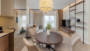 A modern living room with a round wooden table, beige sofas, a large window with curtains, a TV, and a bookshelf. - Cracow Novum Apartment #365 (Kraków)