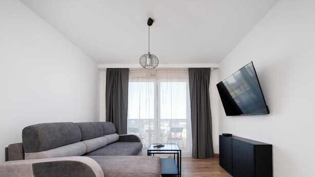 Ein modernes Wohnzimmer mit einem grauen Ecksofa, einem kleinen Tisch, einem an der Wand montierten Flachbildfernseher und einem durch das Fenster sichtbaren Balkon.