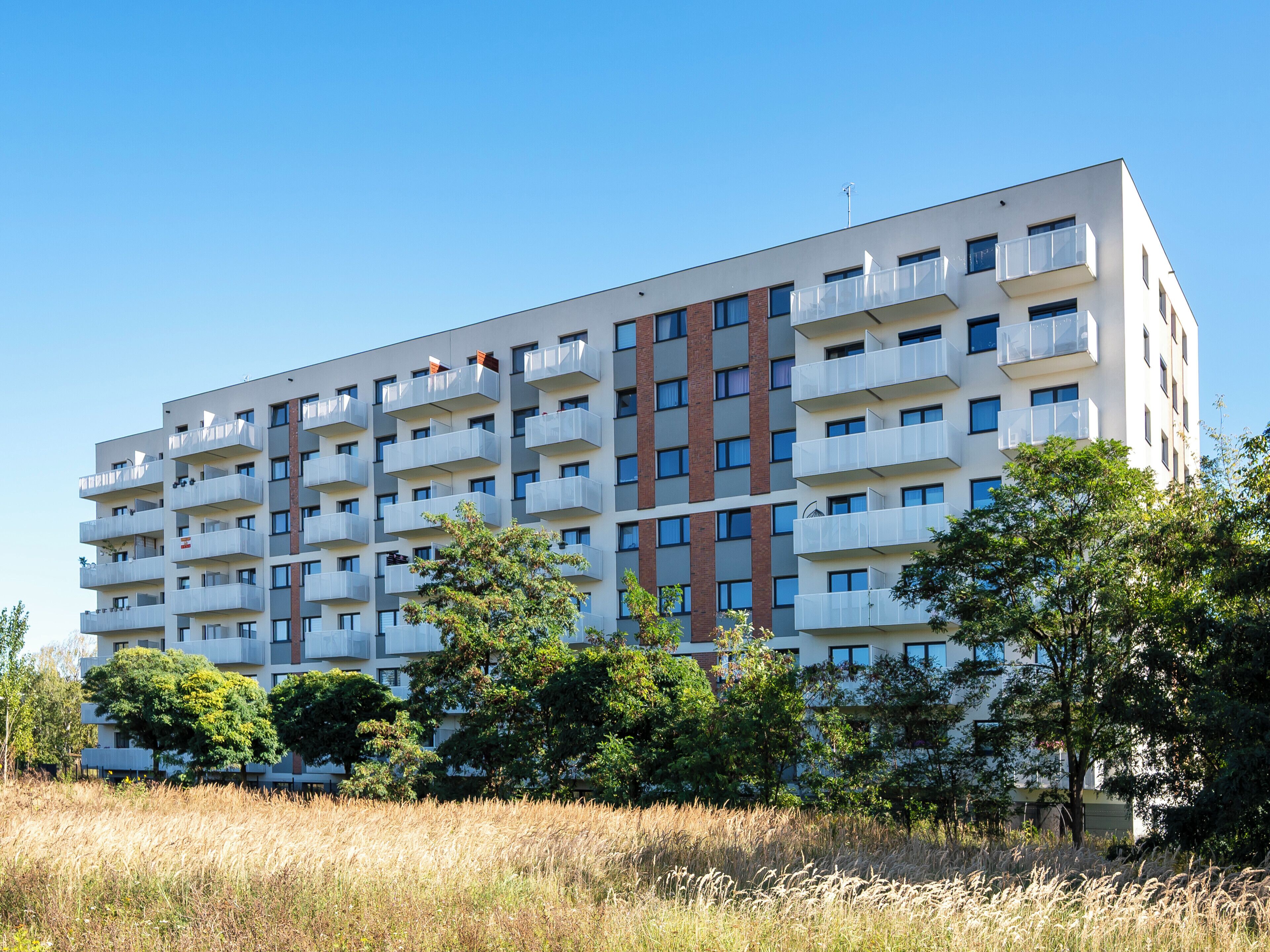 En flervåningsbyggnad med bostäder och balkonger, omgiven av träd och en gräsyta.