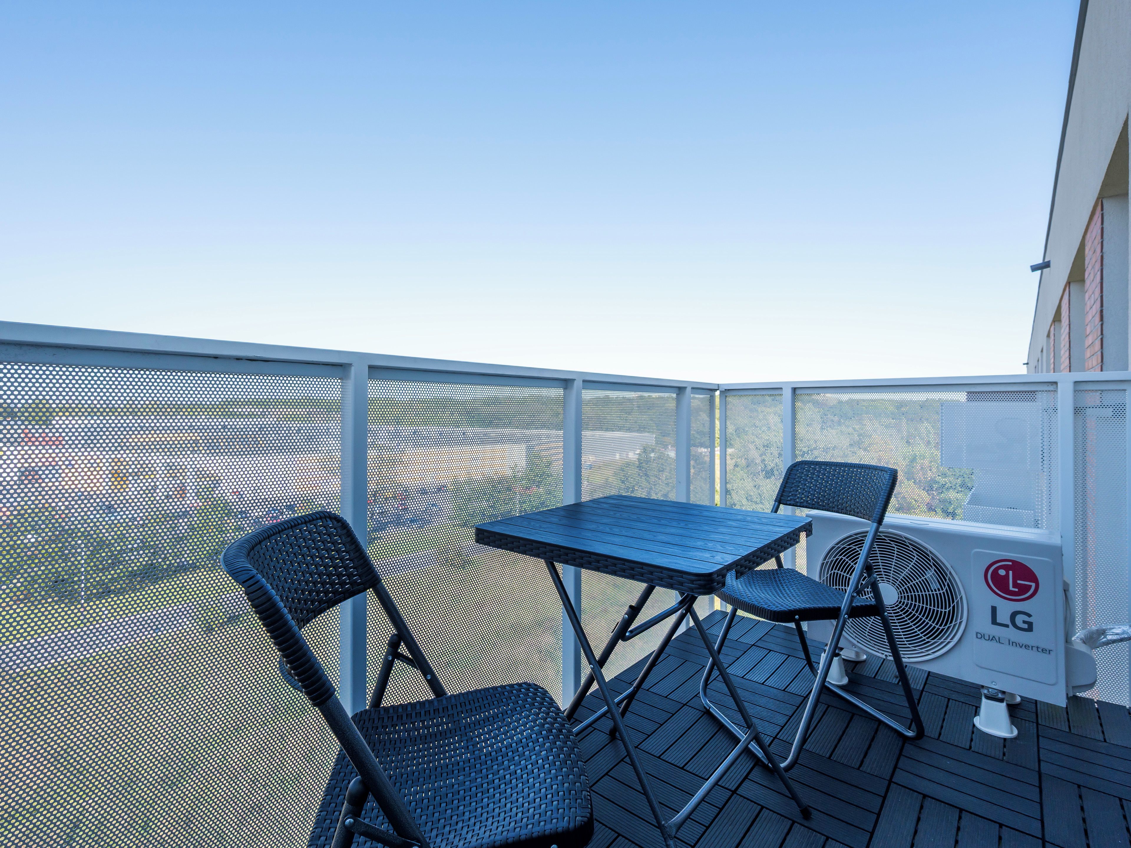 Un balcon extérieur avec une table et deux chaises, un climatiseur LG blanc et une clôture en treillis métallique.