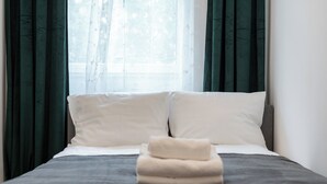 Cama bem arrumada com travesseiros brancos e um cobertor bege dobrado, em frente a cortinas verde escuro e um tecido translúcido na janela.