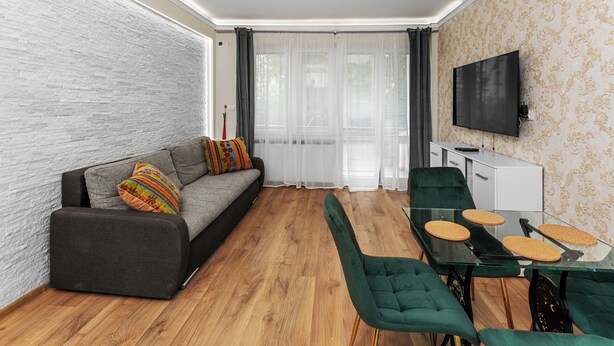 Uma sala de estar moderna com um sofá cinzento, almofadas coloridas, uma mesa de jantar de vidro com cadeiras verdes, uma TV de tela plana e um papel de parede estampado.