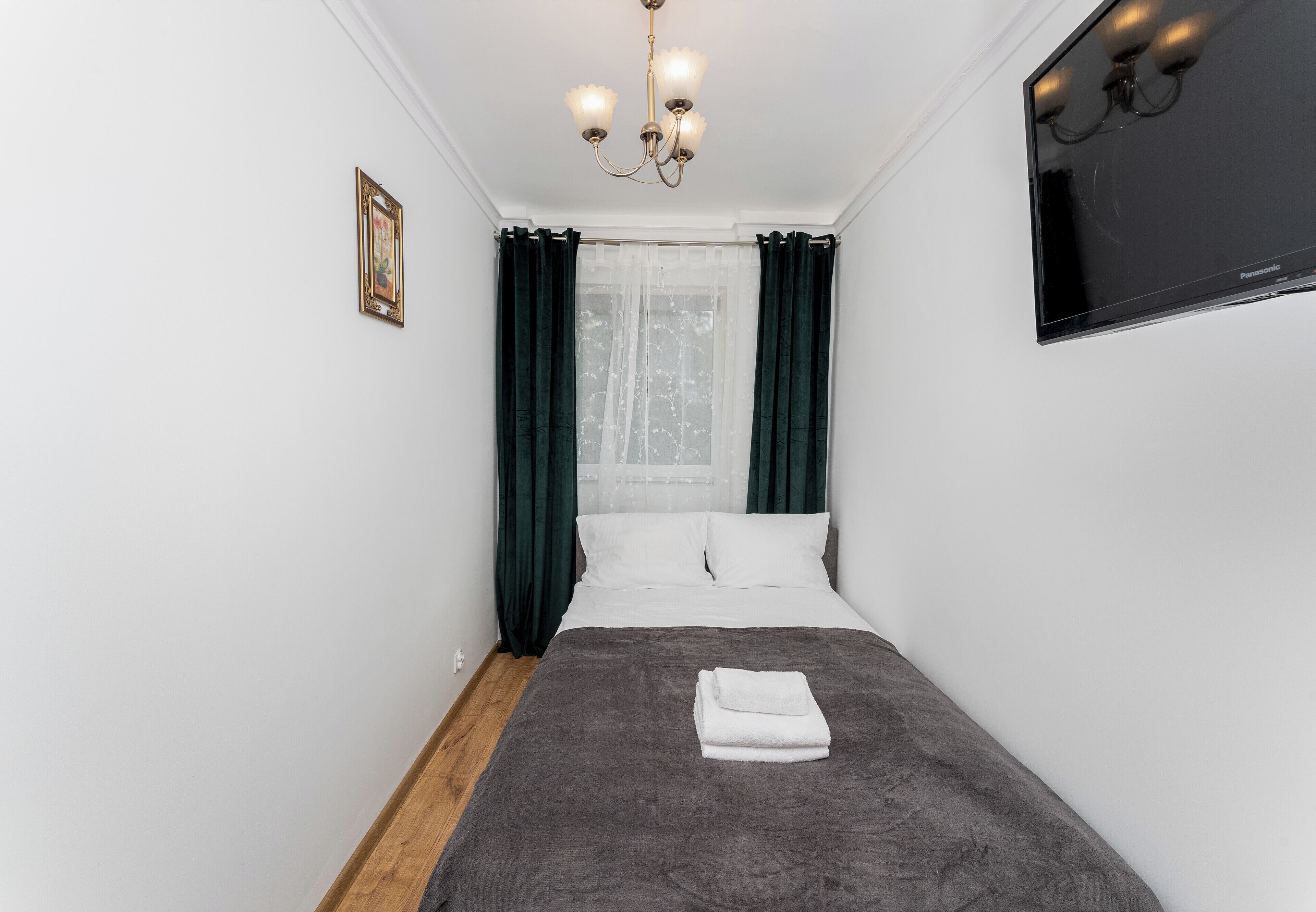 Een net opgemaakt bed met een grijze dekbedovertrek en een gevouwen witte handdoek, een kroonluchter, een ingelijst beeld op de muur en een flatscreen televisie boven het bed.