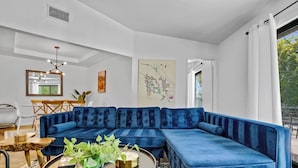 A modern living room with a large blue sectional sofa, a dining area with a wooden table and chairs, and a large window with a view of greenery. - Casa Rafael: Palm Springs Private Pool & Hot Tub (Palm Springs)