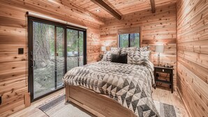 A wooden cabin bedroom with a bed, a nightstand, and a large sliding glass door leading to a forested area. - Songbird at Chambers by Tahoe Getaways - Pet-Friendly 2 BR w/ HOA Beach Pool (Homewood)