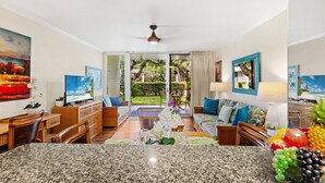 A spacious living room with a large TV, comfortable seating, a kitchen area with granite countertops, and a view of a lush garden through glass doors. - Tropical Charm by Napili Beach (Napili)
