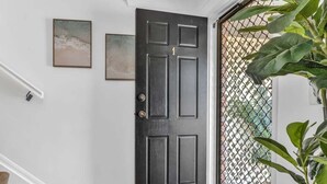 A dark wooden door with a number "1" above it, a white wall with two framed artworks, a wooden staircase with a white handrail, and a large potted plant to the right. - Wave Whisperer - 1/15 Kingscliff (Tweed)