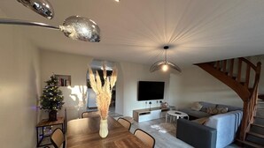 A modern living room with a wooden dining table, a sofa, a TV, and a staircase. - LE MAGNOLIA - LES MOLLETTES (LES MOLLETTES)