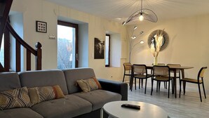 A modern living room with a grey sofa, a dining table with chairs, and a unique ceiling light fixture. - LE MAGNOLIA - LES MOLLETTES (LES MOLLETTES)