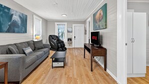 Una sala de estar moderna con un sofá gris, un sillón de masaje, un televisor y suelo de madera.