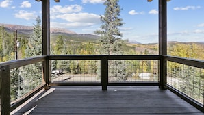 Una terraza de madera con barandilla metálica que da a un paisaje montañoso con árboles y cielo despejado.