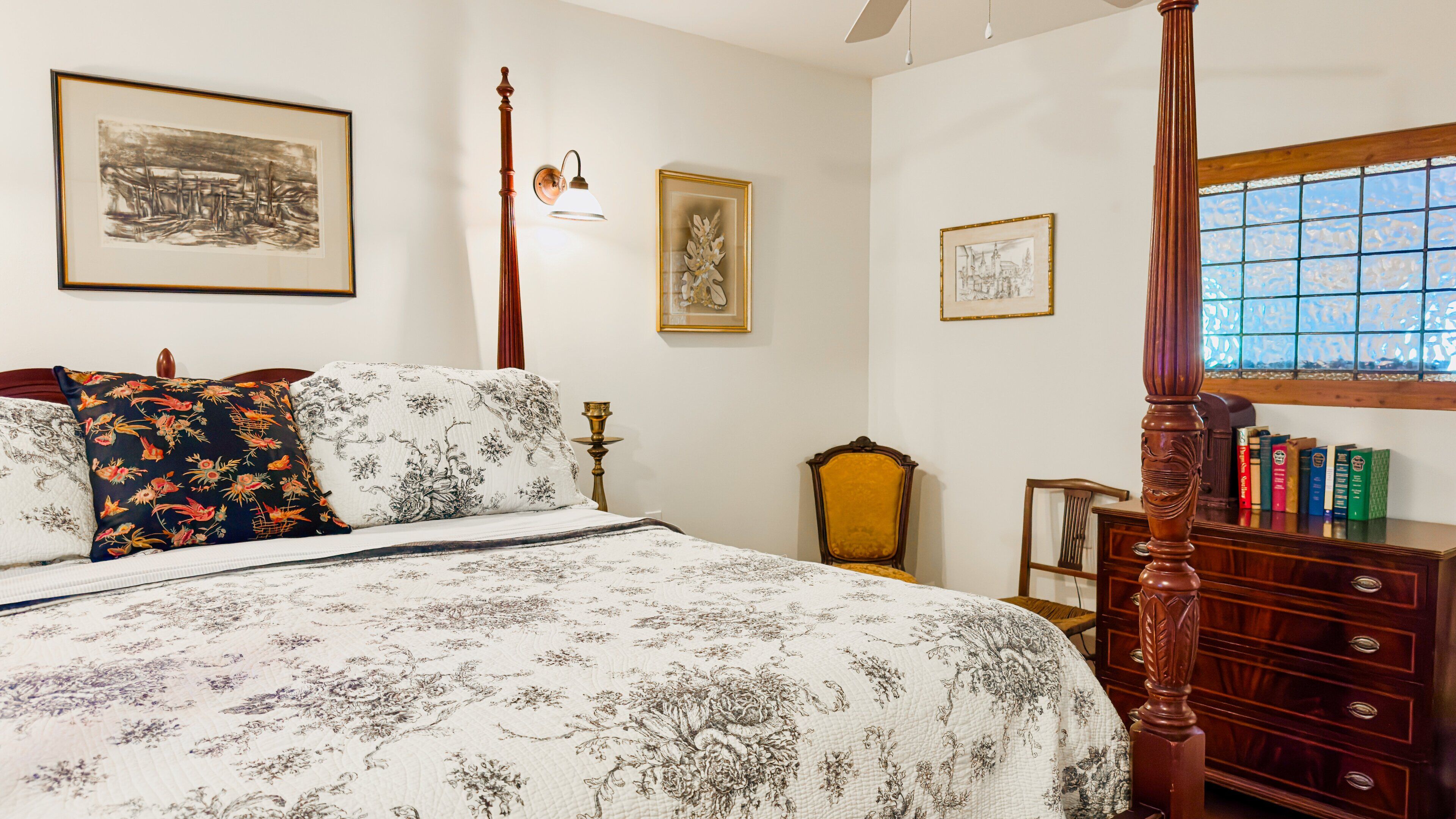 A bedroom with a four-poster bed, floral bedding, a nightstand with books, and a window with a view.
