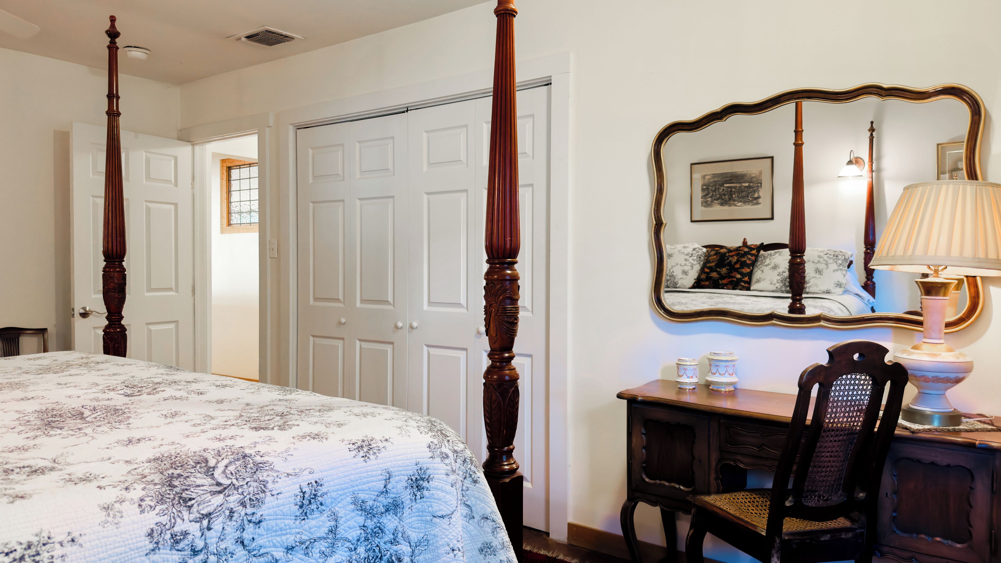A bedroom with a four-poster bed, a desk with a chair, a mirror, and a bedside table with a lamp.