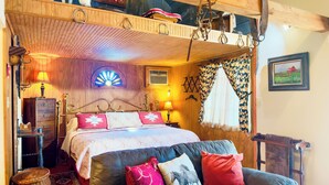 Ein gemütliches Schlafzimmer mit Bett, zwei Nachttischlampen, einer Pendelleuchte, einem Fenster mit Vorhängen, einer Holzbalkendecke und einem Bild an der Wand.