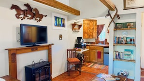 Ein gemütliches Wohnzimmer mit Fernseher, Kamin und Holzmöbeln. Eine Küche mit Holzausstattung und einer Kaffeemaschine ist zu sehen.