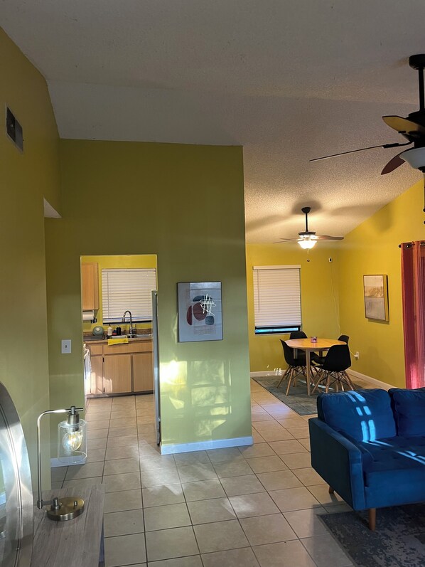 A living room with a blue sofa, a dining area with a table and chairs, a kitchen with a window, and a ceiling fan.