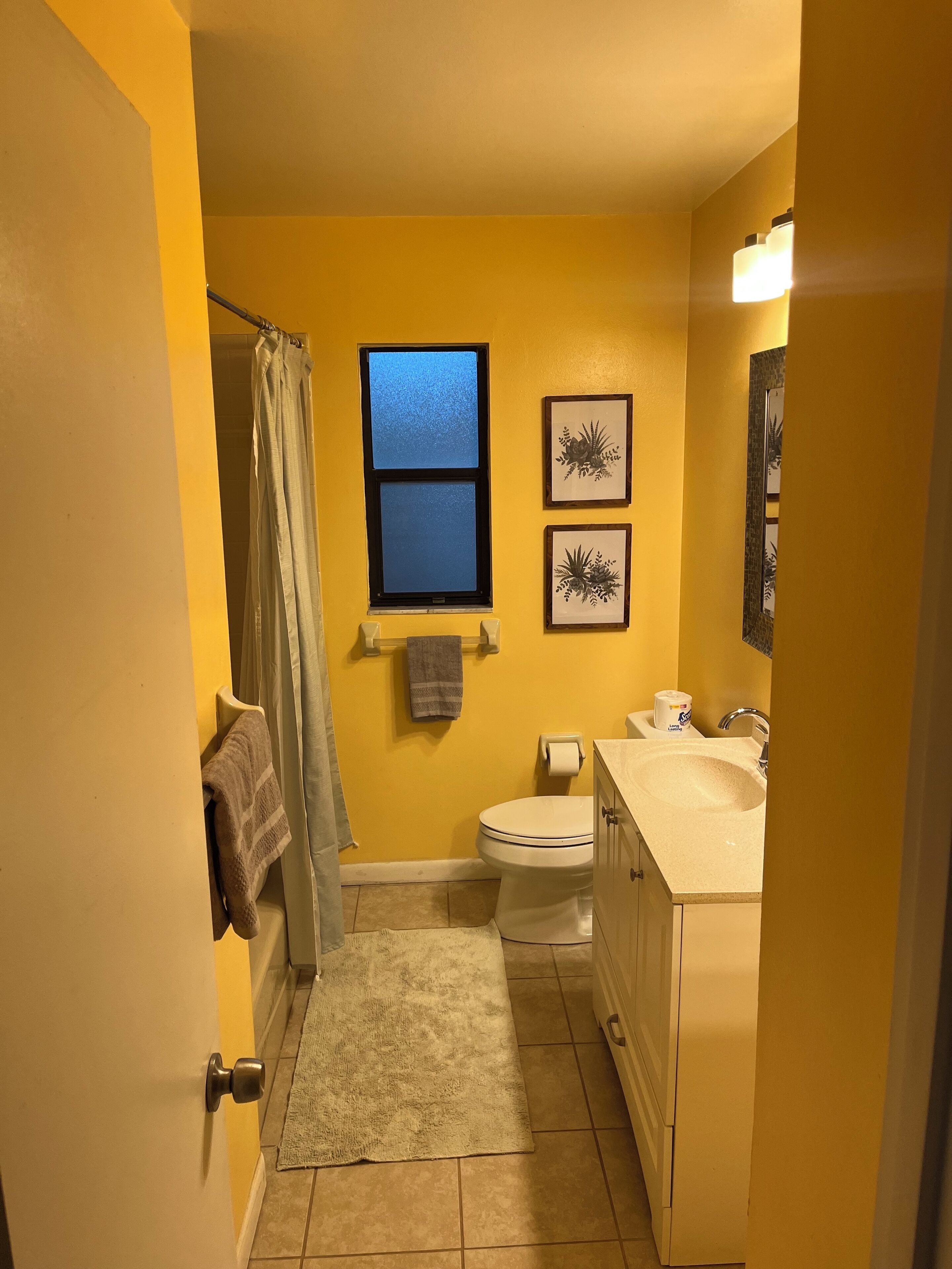 A bathroom with yellow walls, a window, a toilet, a sink, and framed artwork.