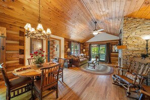 A rustic cabin interior with wooden walls, a dining area with a wooden table and chairs, a cozy living room with a sofa, and a stone wall. - Chic Mountain Cabin with Stream-Lazy River Cabin (Banner Elk)