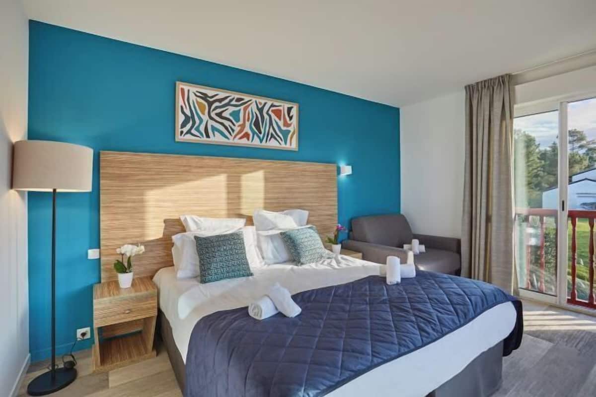 Ein Hotelzimmer mit Bett, Nachttisch, Lampe, Stuhl, Fenster mit Vorhängen und einem Gemälde an der Wand.