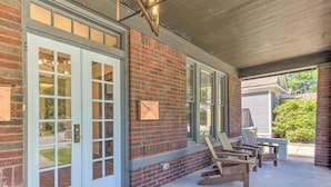 A covered porch with a brick exterior, a white door with glass panels, and two wooden chairs.