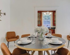 A dining area with a round table set for six, surrounded by six chairs. There is a vase with white flowers on the table, and a window with blinds in the background. - Spacious 3-in-1 Vintage Brick Escape (Memphis)