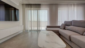 Egy modern nappali, melyben egy nagy, falra szerelt, lapos képernyős televízió, egy bézs színű kanapé, egy márvány kávézóasztal és a padlótól a mennyezetig érő ablakok találhatók, melyekre könnyű, áttetsző függönyök vannak függesztve.