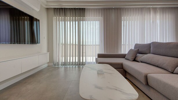 A modern living room with a large flat-screen TV mounted on the wall, a beige sofa, a marble coffee table, and floor-to-ceiling windows with sheer curtains. - Stylish Gozo Penthouse Spacious and Quiet Terrace (Nadur)