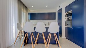 A modern kitchen with dark blue cabinets, a white countertop, and a built-in oven. There are three white chairs with wooden legs. - Stylish Gozo Penthouse Spacious and Quiet Terrace (Nadur)