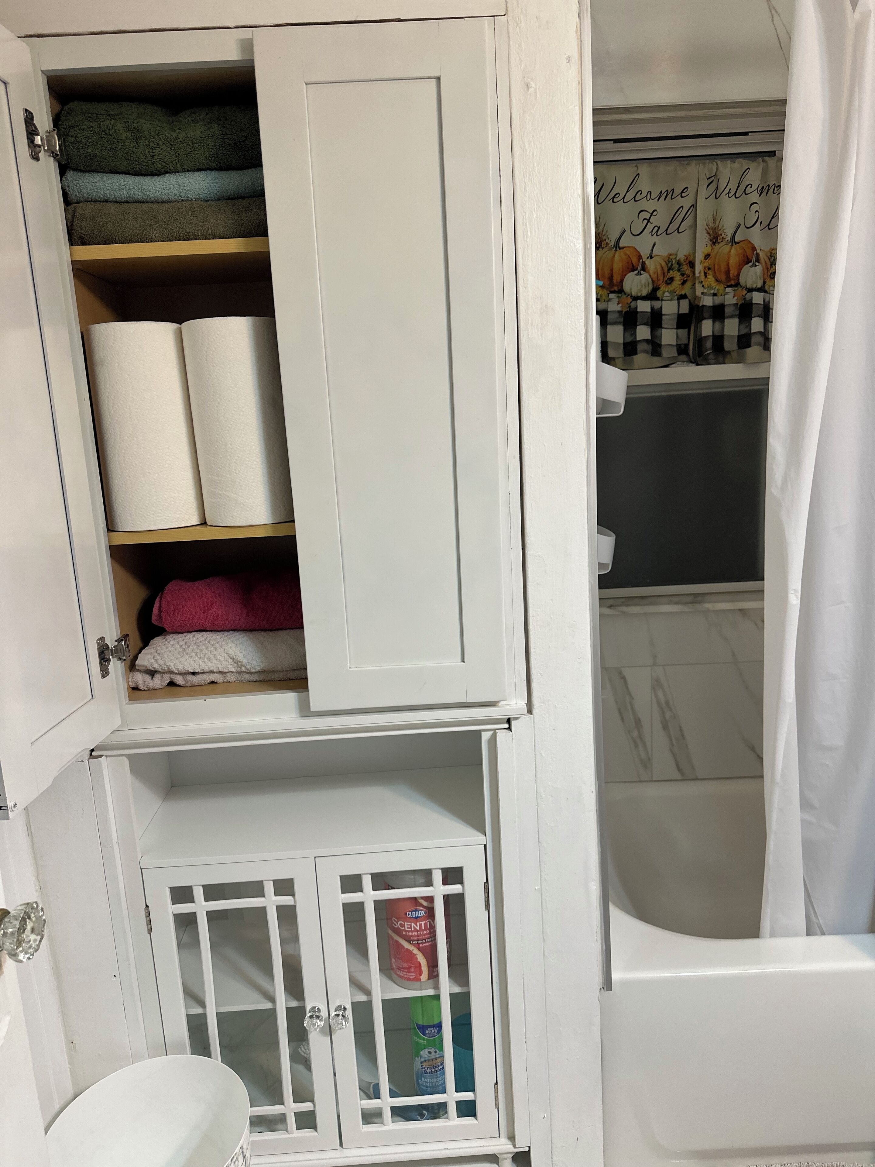 Un baño con un armario blanco que contiene toallas, un inodoro y una cortina de ducha con un cartel que dice 'Welcome Fall'.
